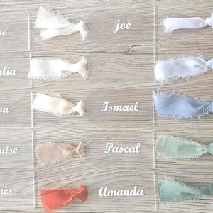 Peut inclure: Une collection d'étiquettes rectangulaires en acrylique transparent aux extrémités arrondies, chacune avec un nom imprimé en blanc et un ruban en tissu effiloché noué. Les noms incluent Julie, Giulia, Eva, Louise, Inès, Joé, Ismaël, Pascal, Amanda, Christopher, Noah, Gabriel, Bilel et Jean-Paul.