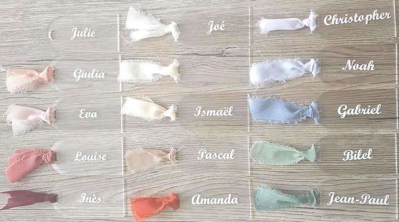 May include: A collection of clear acrylic rectangular tags with rounded ends, each featuring a white printed name and a frayed-edge fabric ribbon tied through a hole. Names include Julie, Giulia, Eva, Louise, In&egrave;s, Jo&eacute;, Isma&euml;l, Pascal, Amanda, Christopher, Noah, Gabriel, Bilel, and Jean-Paul.