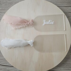 May include: Two rectangular acrylic tags with rounded tops, each featuring a frayed fabric ribbon tied through a hole. One tag displays the name Julie in white script lettering, while the other tag is blank. The tags are clear and transparent.