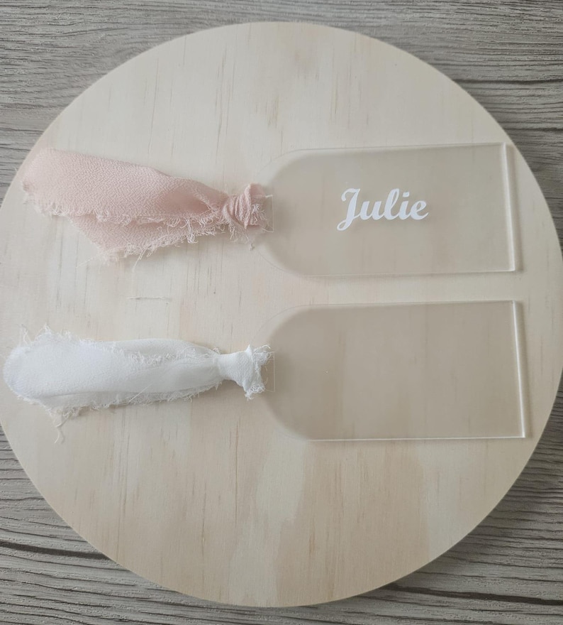May include: Two rectangular acrylic tags with rounded tops, each featuring a frayed fabric ribbon tied through a hole. One tag displays the name Julie in white script lettering, while the other tag is blank. The tags are clear and transparent.