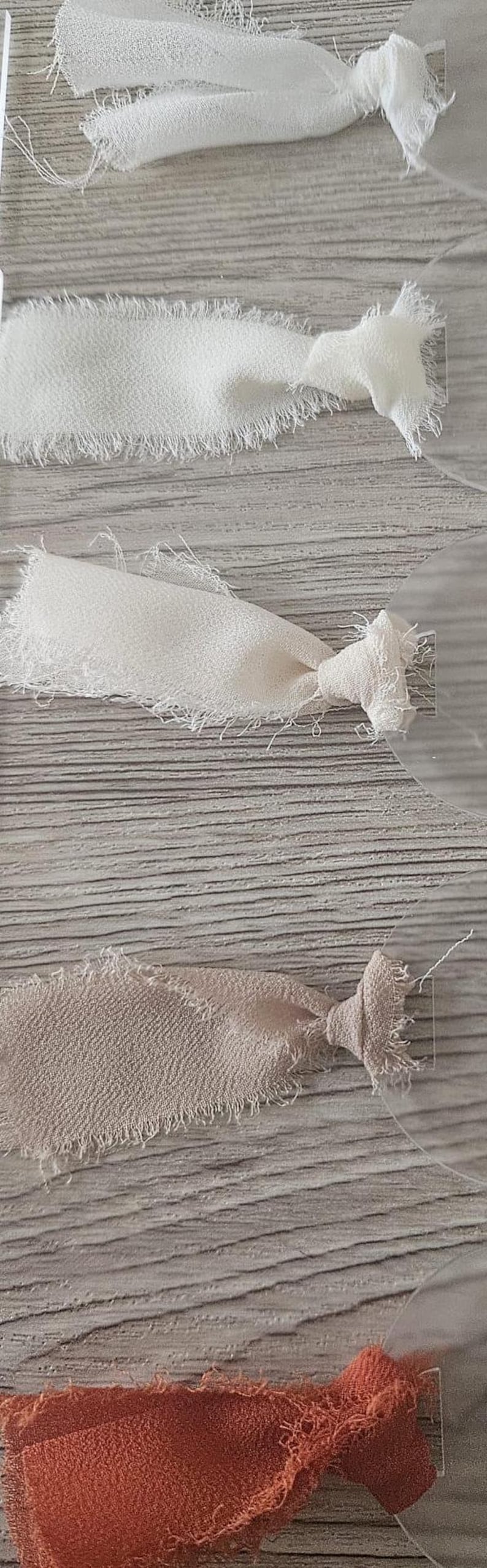 May include: A vertical arrangement of five circular clear acrylic tags, each tied with a strip of frayed-edge fabric in varying shades of white, cream, beige, light tan, and burnt orange, secured with a simple knot through a small hole.