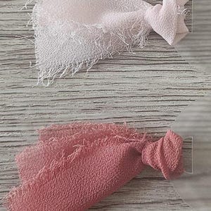 May include: Four fabric swatches with frayed edges attached to circular clear plastic tags. The colors include a pale blush, a light pink, a dusty rose, and a deep burgundy. Each piece of textured fabric is knotted at the base near the tag.