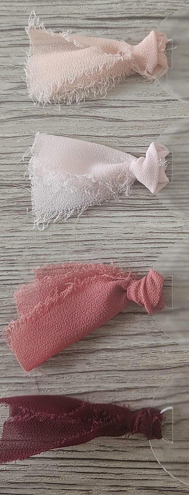 May include: Four fabric swatches with frayed edges attached to circular clear plastic tags. The colors include a pale blush, a light pink, a dusty rose, and a deep burgundy. Each piece of textured fabric is knotted at the base near the tag.