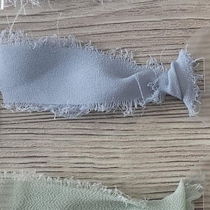 May include: A collection of five fabric ribbons with frayed edges, each attached to a clear acrylic tag. The ribbons are arranged in a vertical gradient, featuring shades of white, light gray, dusty blue, sage green, and dark olive green.