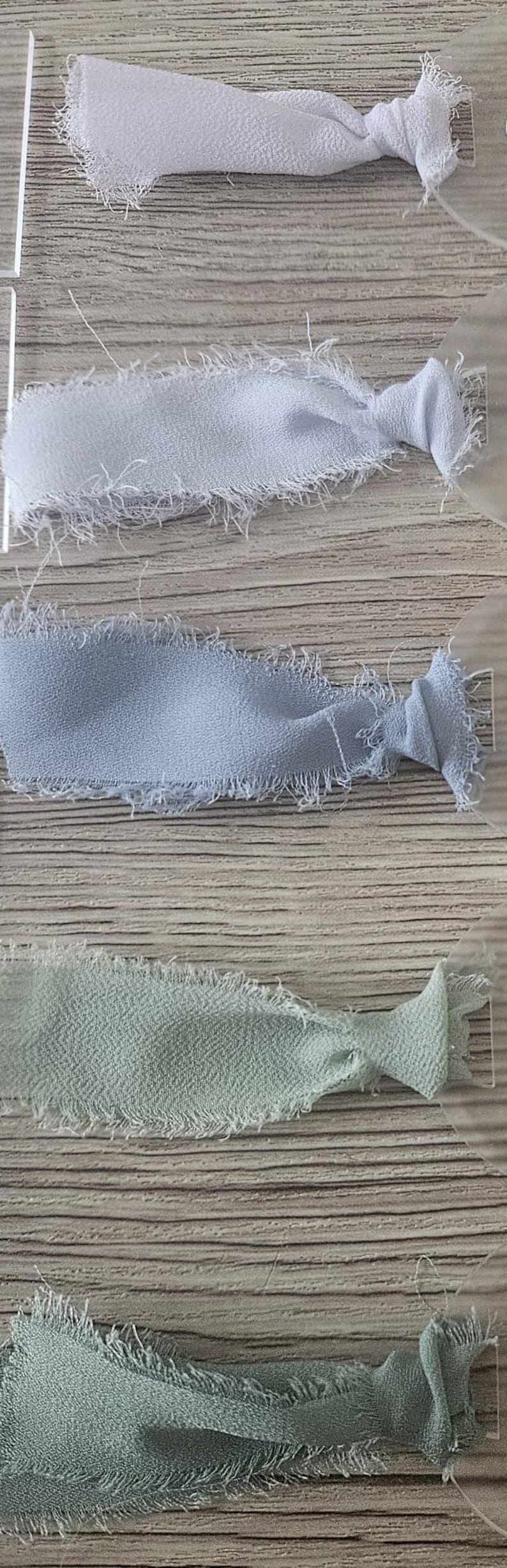 May include: A collection of five fabric ribbons with frayed edges, each attached to a clear acrylic tag. The ribbons are arranged in a vertical gradient, featuring shades of white, light gray, dusty blue, sage green, and dark olive green.