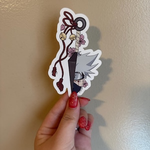 May include: A sticker featuring a split design of a ninja weapon and a character's face. The weapon has a black handle, a dark blade, and a red tassel. The character has gray hair, a mask, and pink flowers.