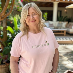 The soft pink Garden Belief t-shirt, worn by a happy woman in her garden.