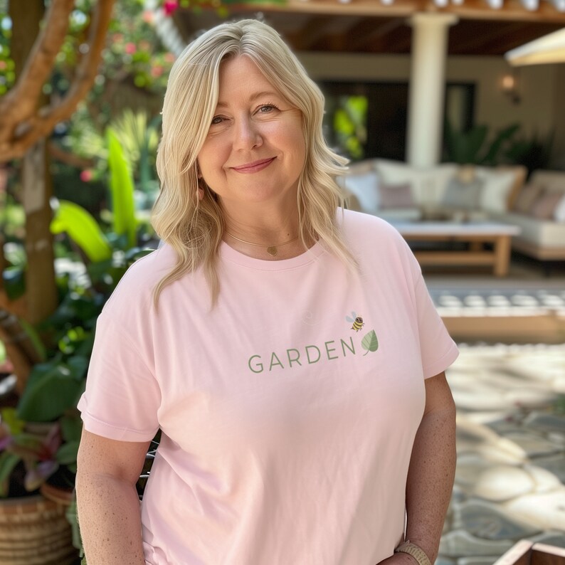 The soft pink Garden Belief t-shirt, worn by a happy woman in her garden.
