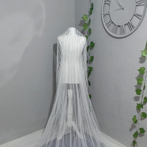 May include: A white bridal veil with a scalloped edge and beaded embellishments. The veil is draped over a mannequin and is shown against a gray background.