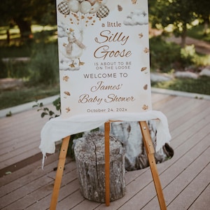 Puede incluir: Un caballete de madera muestra un letrero de baby shower con el texto "a little Silly Goose is about to be on the loose. Welcome to James' Baby Shower." El letrero presenta un ganso de dibujos animados con globos y hojas de otoño.