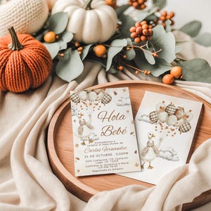 May include: A wooden tray holds two baby shower invitations with a cartoon goose holding balloons. The invitations are decorated with fall-themed elements, including pumpkins and foliage. The text "Hola Bebé" is visible.