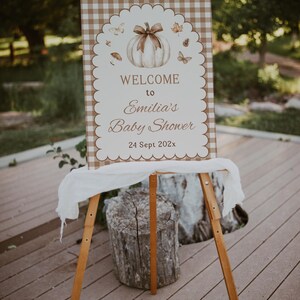 Puede incluir: Caballito de madera que muestra un cartel de bienvenida para un baby shower. El cartel presenta una ilustración de calabaza con un lazo, mariposas y hojas. El texto dice "Welcome to Emilia's Baby Shower, 24 Sept 202x". El cartel tiene un borde festoneado y un patrón de cuadros.