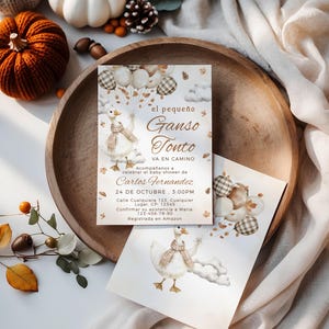 May include: Two baby shower invitations featuring a watercolor illustration of a goose holding balloons. The invitations are on a wooden tray with pumpkins, gourds, and fall foliage. The text on the invitation reads "el pequeño Ganso Tonto".