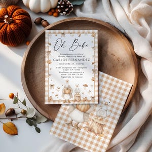 May include: Baby shower invitation with the words "Oh Bebe" in script. The invitation has a tan and white gingham pattern and features illustrations of pumpkins, leaves, and a goose. The invitation is on a wooden tray with pumpkins and fall foliage.