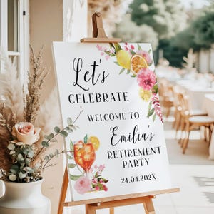 May include: A white sign on a wooden easel with the text "Let's Celebrate Welcome to Emilia's Retirement Party 24.04.202X". The sign is decorated with watercolor illustrations of lemons, oranges, pink flowers, and a cocktail.