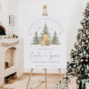 May include: A white sign on a wooden easel with the text "BABIES IT'S COLD OUTSIDE" and "WELCOME TO Emilia & James TWIN BABY SHOWER DECEMBER 25TH. 202X". The sign features a Winnie the Pooh illustration and Christmas trees.