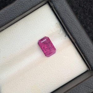 Puede incluir: Una piedra preciosa rosa de forma rectangular, colocada en una caja de presentación blanca.