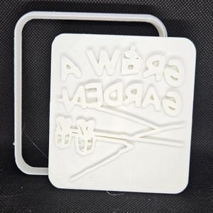 May include: A white, 3D-printed stamp with the text "A Weird Garden" and a stylized flower design. The stamp is square with a rectangular border. The text and design are raised.