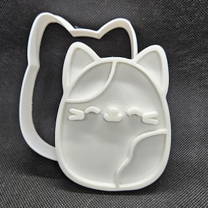 Op de afbeelding: Witte plastic koekjesvormer in de vorm van een cartoonkat met gesloten ogen en een kleine neus. De vormer is ontworpen om koekjes of andere gebakken producten uit te steken.