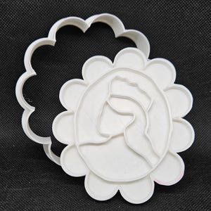 Puede incluir: Cortador de galletas de plástico blanco con forma de flor con la cara de una mujer en el centro. El cortador está abierto, mostrando el interior del diseño.