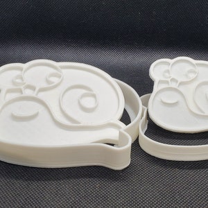 May include: Two white plastic cookie cutters shaped like snails. The larger cutter has a snail with a smaller snail on its back. The smaller cutter is just a single snail.