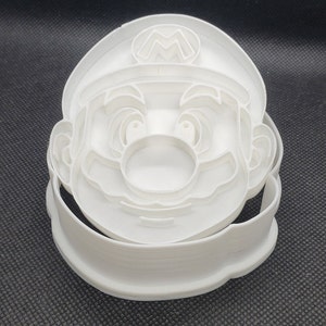 May include: White plastic cookie cutter shaped like the head of the video game character Mario. The cutter has a detailed design of Mario's face, including his mustache, eyes, and hat with the letter "M" on it.