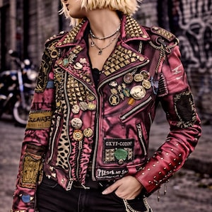 May include: Pink leather jacket with gold studs, patches, and pins. The jacket has a leopard print panel and the text "OXYI-CODIN!" and "PRESTA NO SAL". The jacket is worn with black jeans.