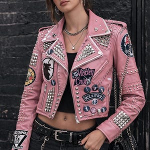 Pode incluir: Jaqueta de couro rosa estilo motociclista com tachas prateadas e patches. Apresenta os textos "Mötley Crüe" e "Rock 'N' Roll". A jaqueta tem um design clássico com gola de lapela e fecho de zíper. Um cinto e uma corrente pretos também são visíveis.