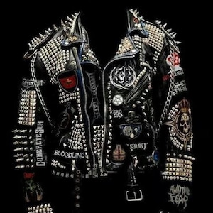Puede incluir: Una chaqueta de cuero negra con tachuelas y pinchos plateados. La chaqueta tiene varios parches, incluyendo un parche de calavera, un parche con el texto "COLD GODS" y un parche con el texto "CONCRETESUN".