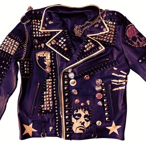Op de afbeelding: Zwarte leren jas versierd met goudkleurige studs, spikes en patches. De jas heeft een ketting, sterren en afbeeldingen van een schedel en een gezicht. De jas heeft een klassieke biker-stijl met ritssluiting en meerdere zakken.