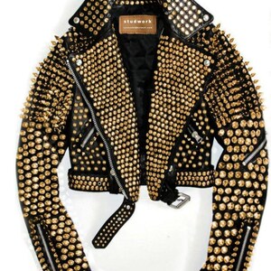 Puede incluir: Chaqueta de cuero negro adornada con tachuelas y púas doradas. Presenta un cuello con solapas, cierre de cremallera asimétrico y mangas con cremallera. La chaqueta tiene un cinturón en la cintura y la marca "studwork" es visible en una pequeña etiqueta.