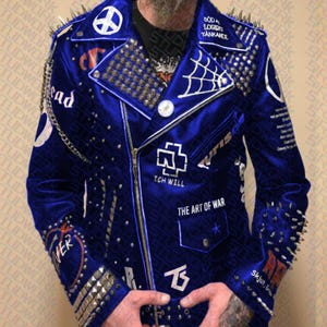 Może przedstawiać: Niebieska aksamitna kurtka ze srebrnymi ćwiekami i kolcami. Kurtka ma białe haftowane wzory, w tym symbol pokoju i pajęczynę. Tekst na kurtce to "Dead", "Ich Will" i "The Art of War".