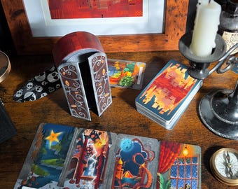 May include: A tarot deck featuring a box with arched doors, a stack of cards with a red and orange city illustration, and four laid-out cards labeled The Star, The Tower, The Moon, and a window scene, alongside a candle holder and a compass.