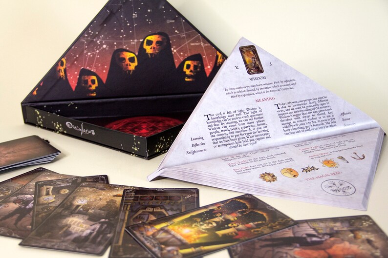 May include: An open triangular box with a celestial design and a set of tarot cards. The box features a dark background with skull-like figures and a red, circular design inside. Several cards are spread out, displaying detailed illustrations.