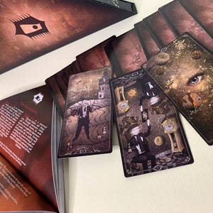 May include: A tarot card deck with a dark, gothic theme. The cards feature detailed illustrations with a vintage aesthetic. The deck includes a box, cards, and a guidebook. The cards depict surreal imagery, including eyes, clocks, and figures in suits.
