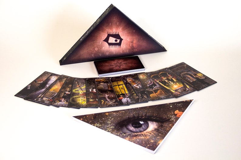 May include: A tarot card deck with a triangular box featuring an eye design. The deck includes a set of cards with detailed illustrations and a smaller rectangular box. The cards are arranged in a fan shape.
