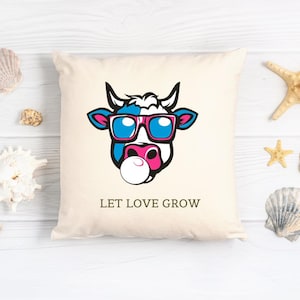 Peut inclure: Coussin carré crème avec un motif de visage de vache de dessin animé. La vache a des détails bleus et roses, des lunettes de soleil et une bulle. Le texte "LET LOVE GROW" est imprimé en dessous. Le coussin est carré.