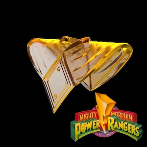 May include: A gold-colored Power Rangers helmet with a clear visor. The helmet is angled, showcasing its design. The Mighty Morphin Power Rangers logo is visible in the lower right corner, featuring a lightning bolt.