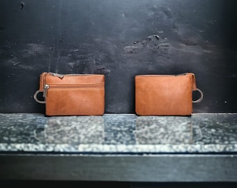May include: Two brown leather zippered pouches with silver zippers and key rings. The pouches are sitting on a gray and white tiled surface.