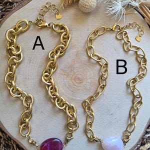 Peut inclure: Deux colliers à chaîne dorée. Le collier A présente une grosse chaîne texturée avec un pendentif en pierre magenta foncé. Le collier B a un design de chaîne similaire avec un pendentif en pierre rose clair. Les deux colliers sont exposés sur une surface en bois.