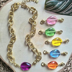 Peut inclure: Un collier en chaîne doré avec une grosse perle violette en forme de goutte d'eau. Cinq perles colorées en forme de goutte d'eau, roses, vertes, jaunes, bleues et oranges, sont également présentées. Les bijoux sont sur une surface en bois.