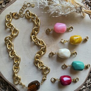Puede incluir: Un collar dorado con un diseño texturizado y una gran cuenta marrón. También se muestran varias cuentas de colores con cierres dorados, incluyendo rosa, amarillo, blanco, turquesa y rojo.