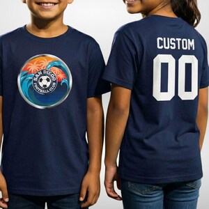 May include: Navy blue t-shirts with a soccer club logo. The front of the shirt features a circular emblem with a soccer ball, palm trees, and ocean waves. The back of one shirt has the word "CUSTOM" and the number "00".
