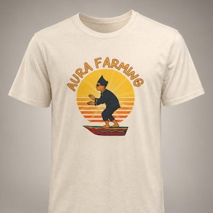 May include: Cream-colored t-shirt featuring a retro design with the text "AURA FARMING" in orange, arched above a figure in a dark outfit, set against a sunset. The figure is standing on a red and brown platform. A casual, everyday wear tee.