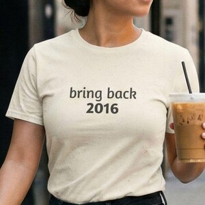 May include: Cream-colored t-shirt with the text "bring back 2016" in black. The person is holding a clear plastic cup filled with iced coffee and a black straw. The shirt is paired with dark denim jeans.