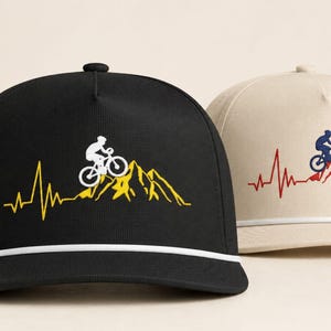 Peut inclure: Une casquette de baseball noire avec un cycliste blanc et un graphique de rythme cardiaque en montagne jaune, et une casquette beige avec un cycliste bleu et un graphique de rythme cardiaque en montagne rouge. Cordon blanc sur la base.