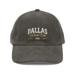 May include: A corduroy baseball cap in a muted olive green. The cap features the text "DALLAS World Cup 2026" in white and gold lettering, with three gold stars above and below the year.