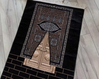 May include: Black prayer rug with a gold and white intricate design featuring a depiction of the Kaaba, the cubical building at the center of Islam's holiest mosque in Mecca, Saudi Arabia.