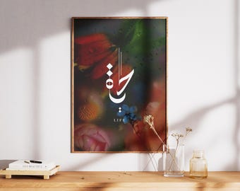 May include: A framed art print featuring a floral arrangement with red, pink, and blue flowers. The artwork includes Arabic calligraphy in white, with the English word "LIFE" below. The print is displayed on a wooden surface.