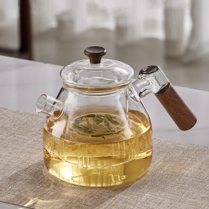 May include: A clear glass teapot with a wooden handle and lid. The teapot is filled with a golden-colored liquid and tea leaves. The handle and lid knob are made of dark wood. The teapot sits on a neutral-colored surface.